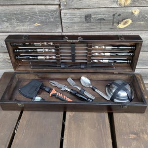 Puede incluir: Una caja de madera con tapa abatible, que contiene un juego de herramientas para asar a la parrilla. Las herramientas incluyen pinchos, un hacha pequeña, una cuchara, un tenedor, un cuchillo y un cuenco de metal. La caja está abierta y las herramientas son visibles.