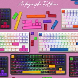 May include: Six different keyboard layouts with rainbow, white, and black keycaps. The keyboards have the text "Autograph Edition" above them. The keyboards are on a pink background with small colorful blocks.