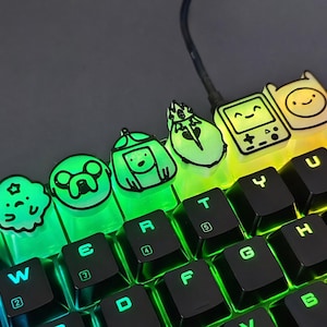 May include: Close-up of a mechanical keyboard with custom keycaps featuring Adventure Time characters. The keycaps are translucent with black outlines and glow with a rainbow backlight. The keyboard is black.