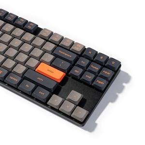 Könnte beinhalten: Eine mechanische Tastatur mit schwarzem Rahmen und grauen und schwarzen Tastenkappen. Die "Enter"-Taste ist orange. Die Tasten haben orangefarbene Schrift. Die Tastatur ist zum Betrachter hin abgewinkelt.