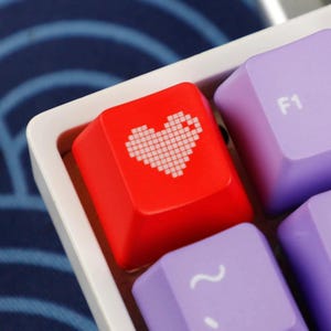 May include: Close-up of a red keyboard key with a white pixelated heart design. Adjacent to the heart key are purple keys, one labeled "F1". The keys are set in a white frame, with a blue patterned background.