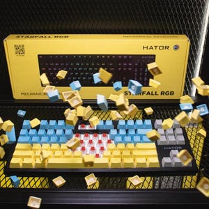 Può includere: Una tastiera meccanica nera con tasti gialli e azzurri. Alcuni tasti sono in aria, come se fossero rimossi. La scatola gialla dietro la tastiera riporta la scritta "STARFALL RGB" e "HATOR".