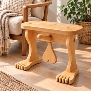 May include: A light-coloured wooden stool featuring a smooth, rectangular seat and two legs shaped like feet with toes. A central horizontal wooden protrusion extends from the frame, with a heart-shaped wooden element hanging below it.