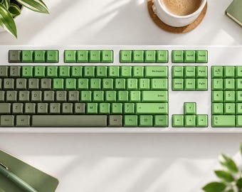 May include: A full-sized computer keyboard with a white frame and a gradient color scheme of keycaps. The keys transition from a dark olive green on the left side to a lighter, bright lime green on the right side, featuring standard QWERTY layout.