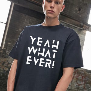 Könnte beinhalten: Ein marineblaues T-Shirt mit dem weißen Text "YEAH WHAT EVER!". Das kurzärmelige Shirt hat eine lockere Passform. Das Bild ist in Innenräumen mit einer Backsteinmauer im Hintergrund aufgenommen.
