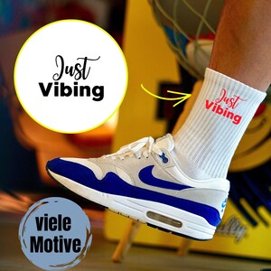 Może przedstawiać: Biało-niebieski but sportowy z białą skarpetą z czerwonym napisem "Just Vibing". But sportowy ma niebieski swoosh i akcenty. Obraz zawiera również tekst "viele Motive" w niebieskim okręgu.