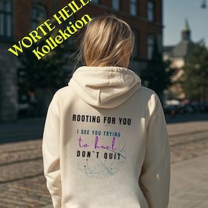 Può includere: Felpa con cappuccio color crema con la scritta "ROOTING FOR YOU, I SEE YOU TRYING to heal, DON'T QUIT" in nero e verde acqua. La felpa con cappuccio ha anche una grafica di linee interconnesse. Le parole "WORTE HEILEN Kollektion" sono in giallo in alto.