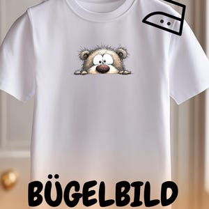 May include: White t-shirt with a cartoon bear design. The bear has large eyes, a brown nose, and light brown fur. The shirt has the text "BÜGELBILD" at the bottom and an iron symbol.