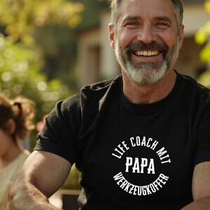 Könnte beinhalten: Ein schwarzes T-Shirt mit dem Text "LIFE COACH MIT PAPA WERKZEUGKOFFER" in einem weißen kreisförmigen Design. Das Bild zeigt eine lächelnde Person, die das T-Shirt trägt.
