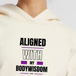 Könnte beinhalten: Cremefarbener Kapuzenpullover mit dem Text "ALIGNED WITH MY BODYWISDOM" in Schwarz und Lila. Der Text ist mittig auf der Vorderseite platziert. Der Hoodie hat eine Kapuze und einen V-Ausschnitt.