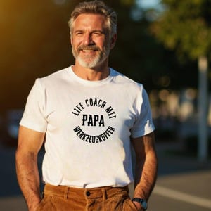 Könnte beinhalten: Ein weißes T-Shirt mit dem Text "LIFE COACH MIT PAPA WERKZEUGKOFFER" in einem schwarzen kreisförmigen Design. Die Person trägt eine braune Cordhose und eine schwarze Uhr. Das Bild ist im Freien aufgenommen.