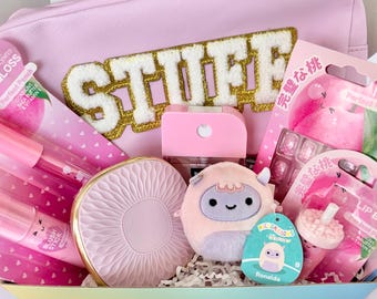May include: A light pink pouch with the word STUFF in gold and white letters, containing a collection of beauty items including lip gloss, a blush stick, a compact mirror, a small plush toy named Ronalda, and a lip balm in a boba tea cup shape.