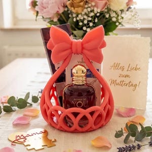 May include: A coral-coloured silicone basket with a large bow handle containing a dark glass perfume bottle labelled Power of You, a rectangular box, and a cream-coloured card with the text Alles Liebe zum Muttertag, plus a gold puzzle piece labelled Mama.