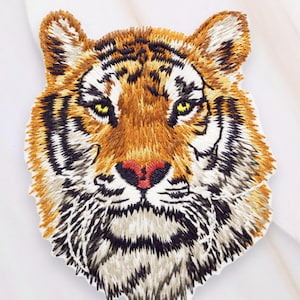Peut inclure: Écusson brodé tête de tigre avec fil orange, marron, noir et blanc. Le tigre a des yeux jaunes et un nez rouge. Une pièce d'art textile détaillée.