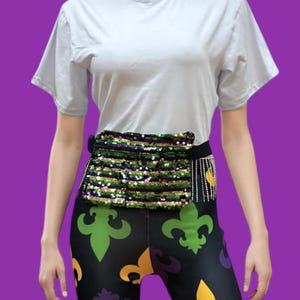 May include: A sequined fanny pack with green, gold, and black sequins is worn around the waist. The fanny pack is paired with black leggings featuring a fleur-de-lis pattern in green, gold, and purple. A white t-shirt completes the outfit.