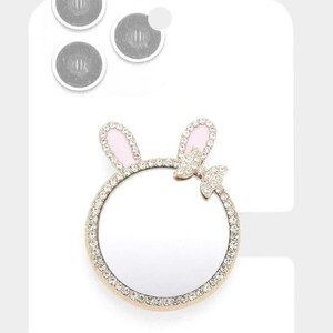 May include: A gold-toned phone ring holder with a bunny ear design. The ears are pink with a crystal border, and the ring is encrusted with crystals. A small crystal bow is at the base of the ears. Includes three adhesive pads.