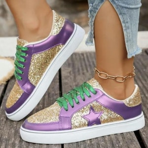 May include: A pair of glittery sneakers with gold and purple accents. The sneakers have a white sole, green laces, and a star design on the side. The shoes are on a wooden surface.