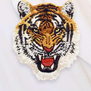 Op de afbeelding: Geborduurde tijgerkop-patch met oranje, zwarte en witte details. De bek van de tijger staat open en onthult witte tanden en een rode tong. De patch staat op een witte achtergrond.