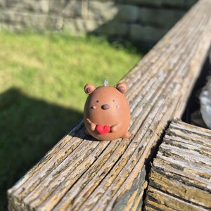 May include: A brown bear-shaped ornament holding a red heart. The bear has small, round ears and a silver loop for hanging. The ornament is made of a smooth, clay-like material.