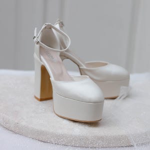 May include: A pair of white satin platform heels with ankle straps. The heels are approximately 4 inches tall and have a platform that is about 1 inch thick.