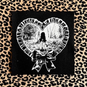 May include: Black square patch with a white heart-shaped design depicting a forest scene and a figure. The heart is framed with a ruffled edge and a bow. The background is a leopard print.