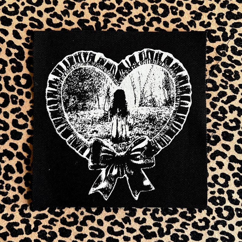 May include: Black square patch with a white heart-shaped design depicting a forest scene and a figure. The heart is framed with a ruffled edge and a bow. The background is a leopard print.