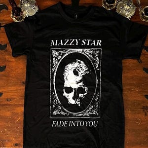 Könnte beinhalten: Schwarzes T-Shirt mit dem Schriftzug "MAZZY STAR" über einer weißen Totenkopf-Grafik in einem ovalen Rahmen. Darunter steht "FADE INTO YOU". Das T-Shirt liegt auf einer Holzoberfläche.
