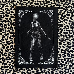 May include: Black rectangular patch with a white illustration of a figure holding a knife, framed by a decorative border. The figure is in a standing pose. The background is black.