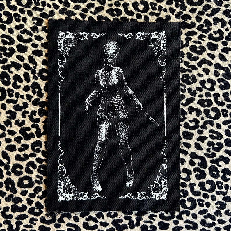 May include: Black rectangular patch with a white illustration of a figure holding a knife, framed by a decorative border. The figure is in a standing pose. The background is black.