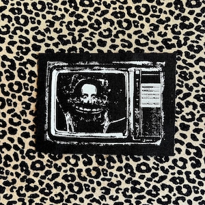 May include: A black patch depicting a retro television set with a distorted face on the screen. The image is in monochrome, with a vintage aesthetic. The patch is rectangular and has a stitched border.