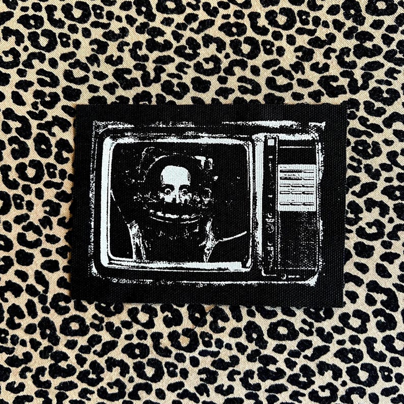 May include: A black patch depicting a retro television set with a distorted face on the screen. The image is in monochrome, with a vintage aesthetic. The patch is rectangular and has a stitched border.