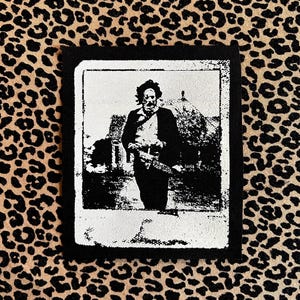 May include: Black and white patch featuring a vintage image of a man with a chainsaw. The image is framed in a white border with a black background. The patch is set against a leopard print background.