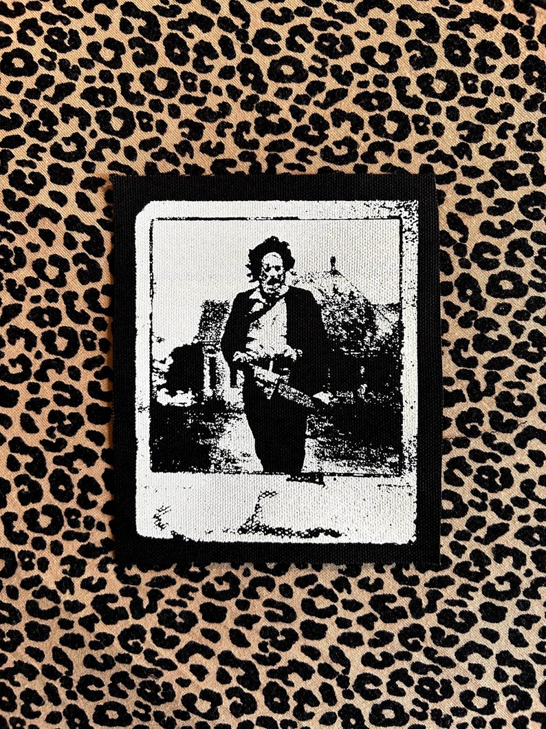 May include: Black and white patch featuring a vintage image of a man with a chainsaw. The image is framed in a white border with a black background. The patch is set against a leopard print background.
