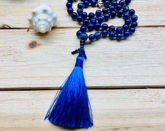 May include: A blue lapis lazuli mala with silver accents and a blue tassel. The mala is laid out on a wooden surface.