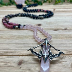 May include: A pink crystal pendant necklace with a silver-colored crossbow design. The necklace features pink and black beaded strands. A rose quartz crystal is visible in the background.