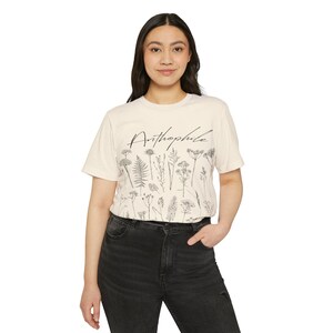 May include: A cream-colored t-shirt featuring a botanical illustration of various plants and the word "Anthophile" in a script font. The design is in black and white. The person is wearing black jeans.