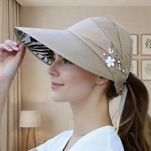 May include: A beige sun visor hat featuring a wide brim with a black and white botanical leaf print on the underside. The exterior side of the visor is adorned with small white flower-shaped embellishments and silver-toned studs.