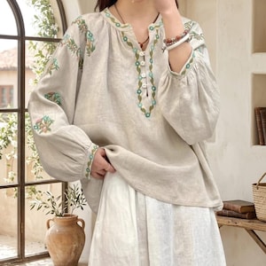 Op de afbeelding: Beige linnen blouse met bloemenborduurwerk in blauw en groen. De blouse heeft een V-hals, knoopdetails en lange, gepofte mouwen. De outfit bevat een witte rok, wat een bohemien esthetiek creëert.