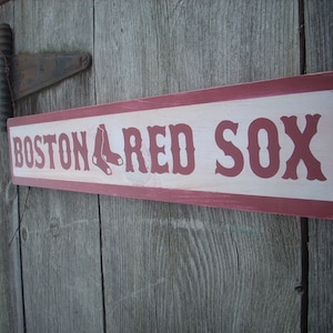 May include: A wooden sign with red and white paint. The sign reads "Boston Red Sox" in red letters.