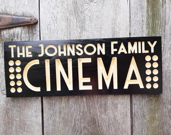 以下が含まれることがあります： 黒い木製の看板に、白い文字で「The Johnson Family Cinema」と書かれています。 「Cinema」という文字は、大きくスタイリッシュなフォントで書かれており、両側に小さな円が並んでいます。
