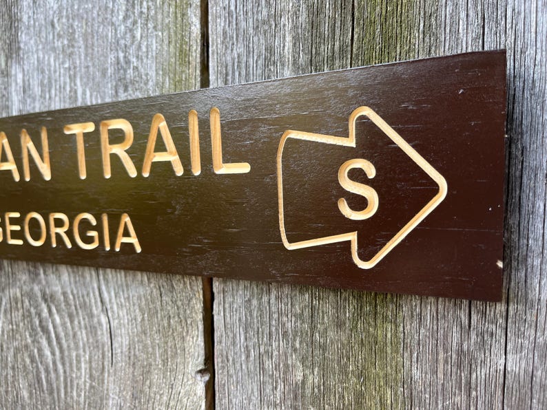 May include: A dark brown wooden sign with the words "Appalachian Trail Georgia" and an arrow pointing to the right with the letter "S" inside.