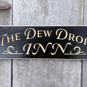 May include: A rectangular black wooden sign with the words "The Dew Drop Inn" carved in a light beige color. The sign has decorative flourishes and is mounted on a weathered wooden door.