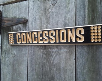 以下が含まれることがあります： 黒い木製の看板に、明るい木で「CONCESSIONS」という文字が彫られています。看板の両端には、小さな丸い明るい木製の円が並んでいます。