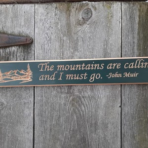 Könnte beinhalten: Ein dunkelgrünes Holzschild mit einer geschnitzten Bergszene und dem Zitat "Die Berge rufen und ich muss gehen. -John Muir" in kupfernen Lettern.