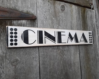 以下が含まれることがあります： 「CINEMA」と黒文字で書かれた白い木製の看板。看板の文字の両側に黒と白の幾何学模様がある。