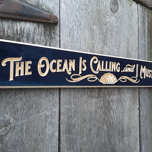 May include: A wooden sign with a dark blue background and gold lettering. The sign reads "The Ocean Is Calling and I Must Go" with a decorative seashell design.