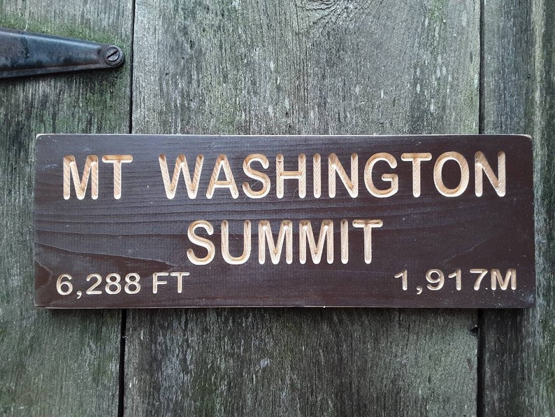 May include: A brown wooden sign with white lettering that reads "MT WASHINGTON SUMMIT" with the elevation listed as "6,288 FT" and "1,917M".