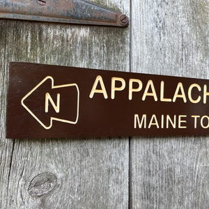 May include: A brown wooden sign with white text that reads "Appalachian Trail Maine to Georgia" and a white arrow pointing left with the letter "N" inside.