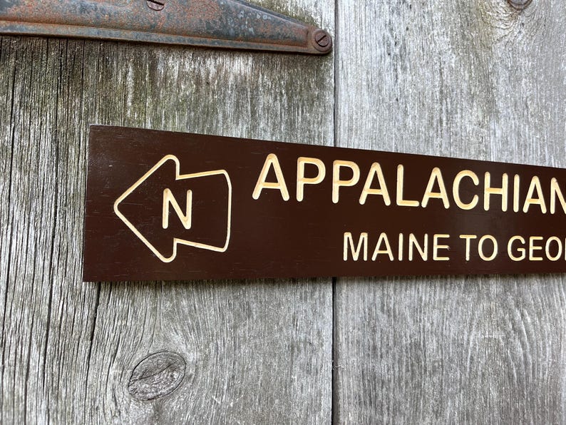 May include: A brown wooden sign with white text that reads "Appalachian Trail Maine to Georgia" and a white arrow pointing left with the letter "N" inside.