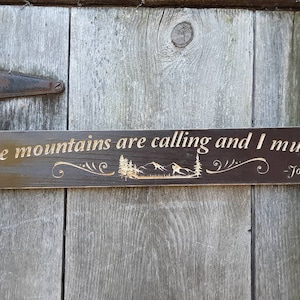 Könnte beinhalten: Ein dunkelbraunes Holzschild mit dem Text "The mountains are calling and I must go. - John Muir" in goldenen Buchstaben. Das Schild zeigt ein stilisiertes Bild von Bergen und Bäumen.